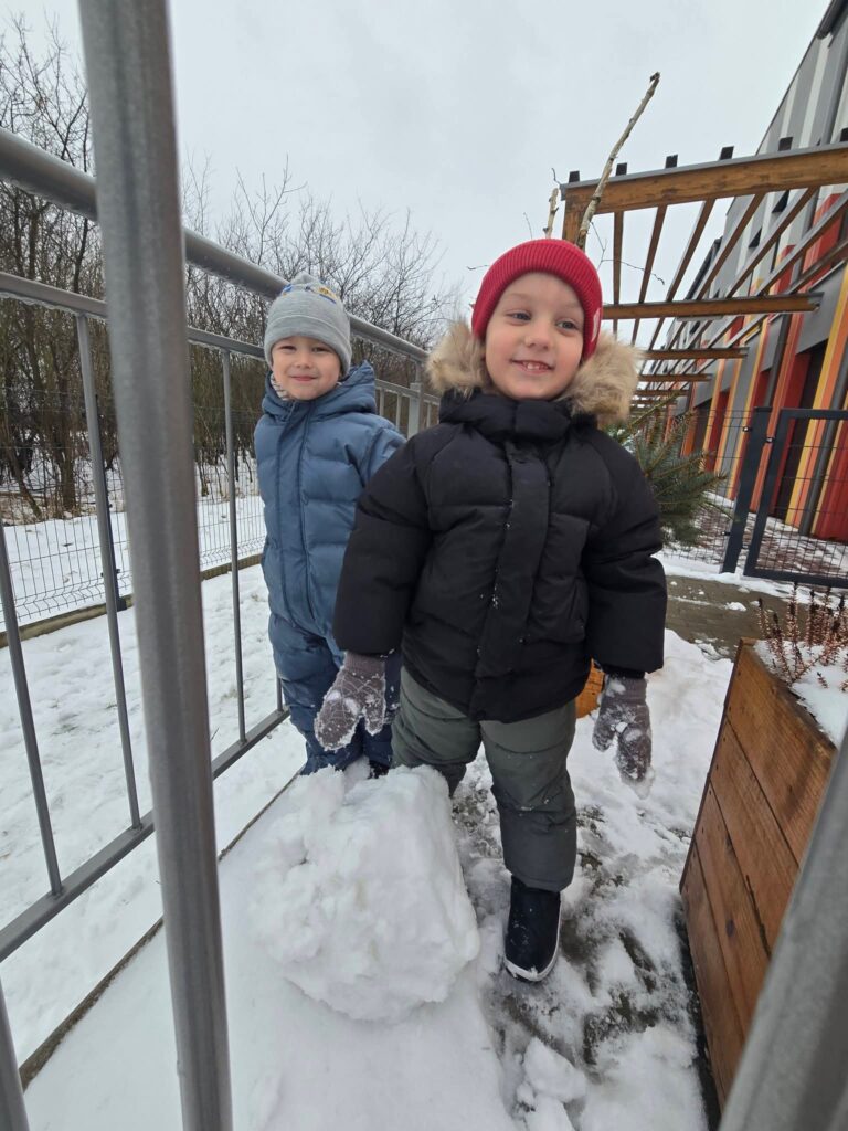 Dzieci ubrane w kolorowe kombinezony lepią duże kule śniegowe na bałwana.