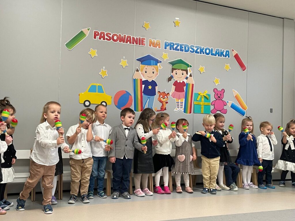 Dzieci stoją w rzędzie, śpiewają i grają na grzechotkach.