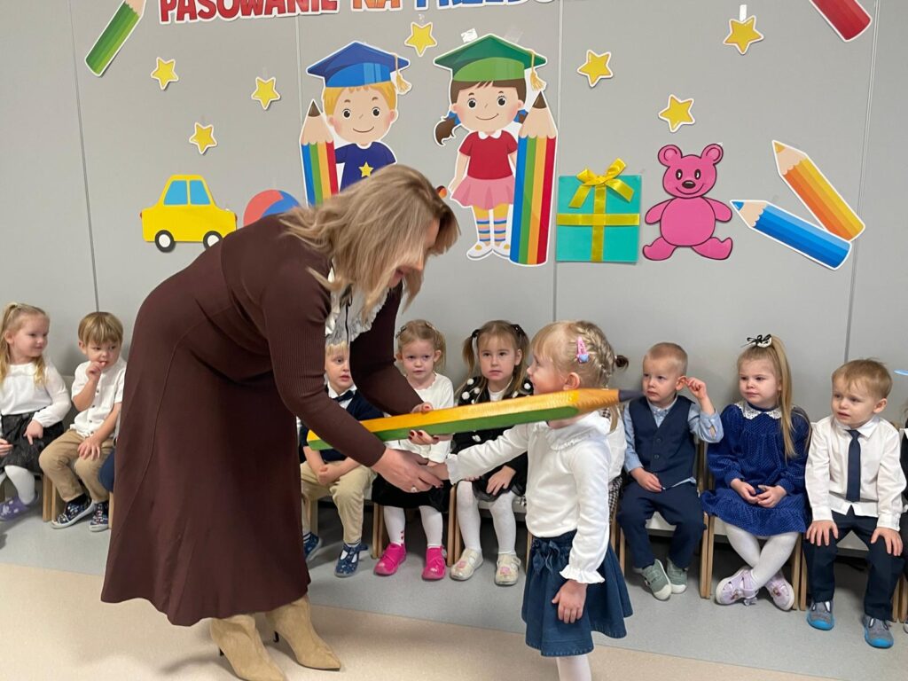 Pani Dyrektor pasuje dziewczynkę na Przedszkolaka. W tle siedzą dzieci.