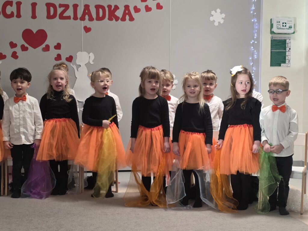 Grupka dzieci ubranych w stroje biało-czarno-pomarańczowe w rękach trzyma kolorowe chustki i śpiewa piosenkę. Za nimi szara ściana z czerwonymi serduszkami.
