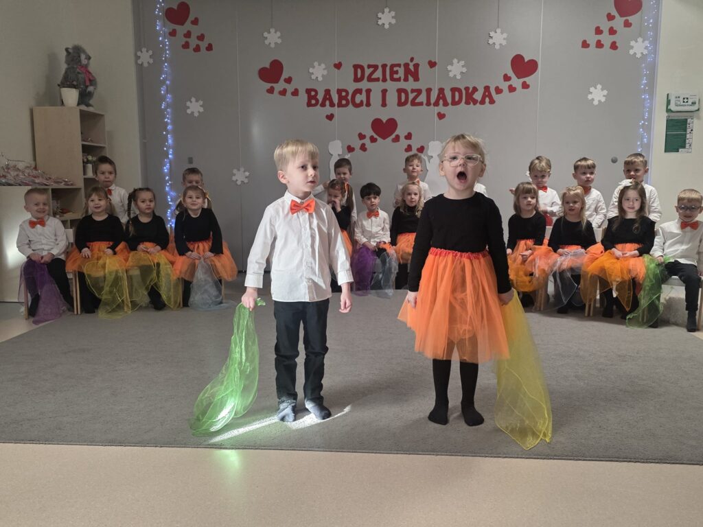 Chłopiec i dziewczynka ubrani w biało-czarno-pomarańczowe stroje, stoją na szarym dywanie i witają gości. W rękach trzymają kolorowe chusteczki.