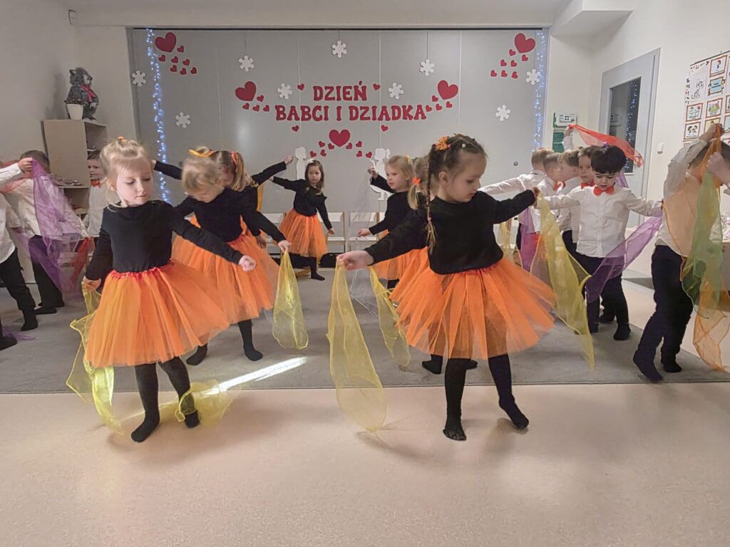 Grupa dziewczynek ubrane na czarno w pomarańczowe spódniczki tańczą na szarym dywanie.