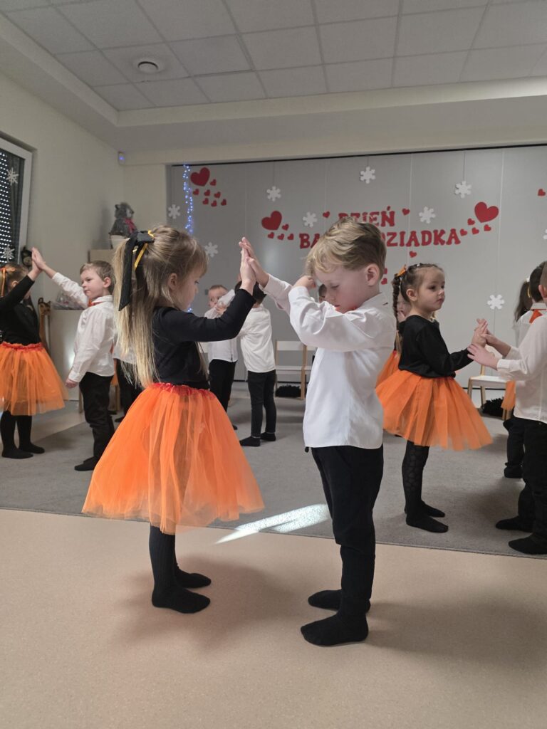 Chłopcy i dziewczynki ubrani w biało-czarno-pomarańczowe stroje, stoją na szarym dywanie i tańczą do muzyki.