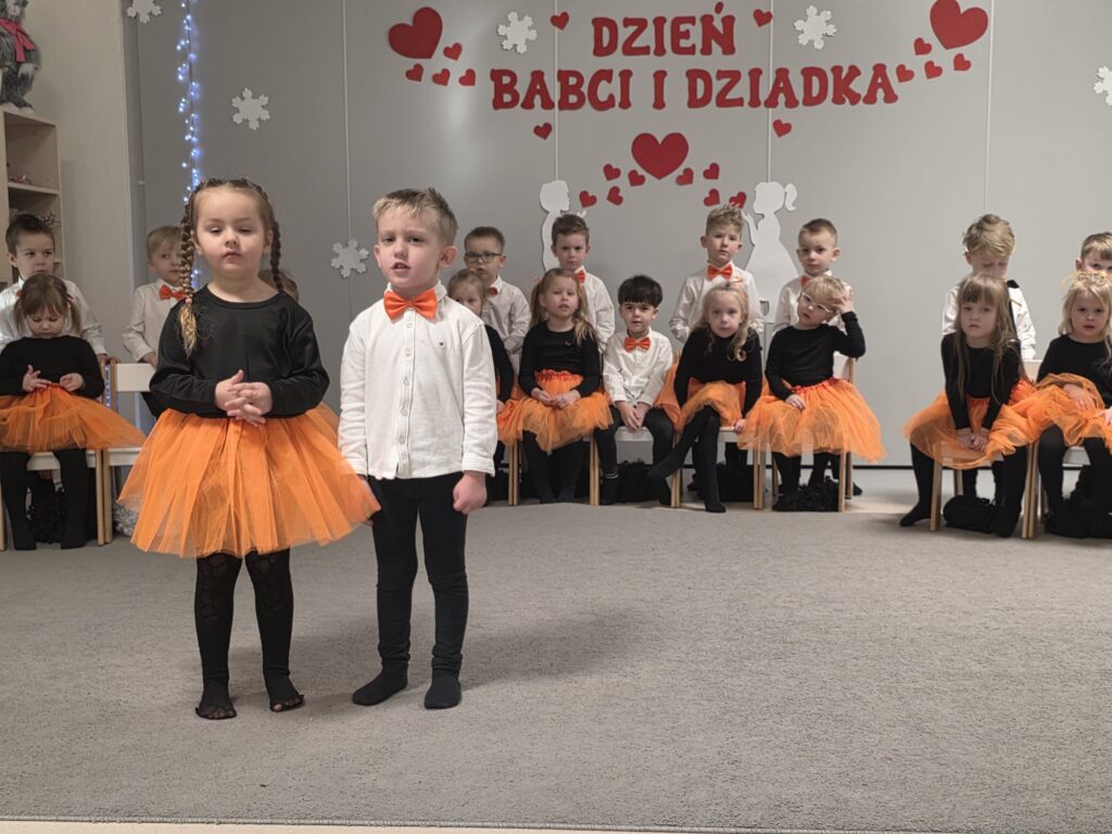 Chłopiec i dziewczynka ubrani w biało-czarno-pomarańczowe stroje, stoją na szarym dywanie i recytują wiersz.