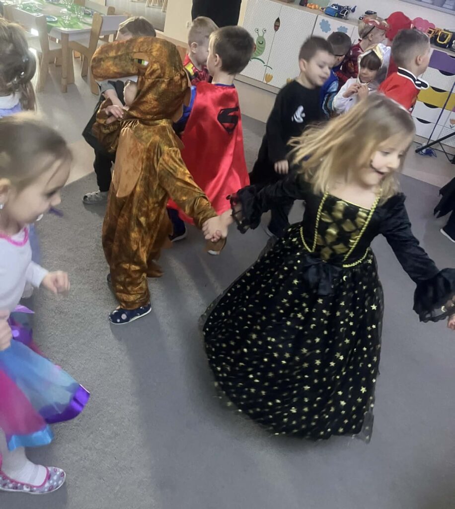 dzieci przebrane w stroje karnawałowe tańczą na dywanie, dziewczynka w czarnej sukience trzyma się za ręce z dinozaurem