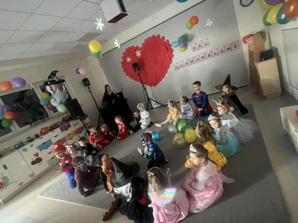 dzieci przebrane w stroje karnawałowe siedzą na dywanie, po środku są balony, z tyłu pani dj