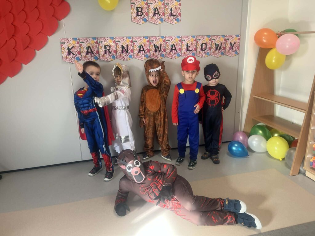 chłopcy stoją przy napisie bal karnawałowy, są przebrani w stroje karnawałowe: superman, kosmonauta, dinozaur, mario, spiderman, i jeleń