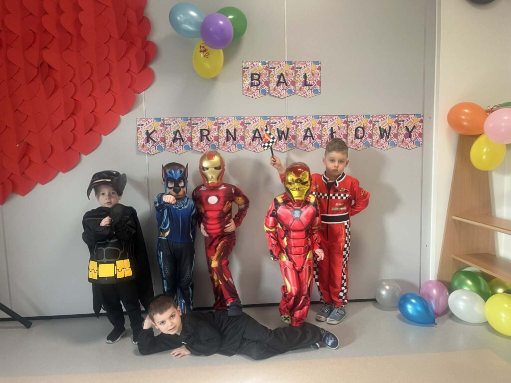 chłopcy stoją przy napisie bal karnawałowy, chłopcy są przebrani w stroje karnawałowe: batman, ironman, kierowca rajdowy, ninja, i pies