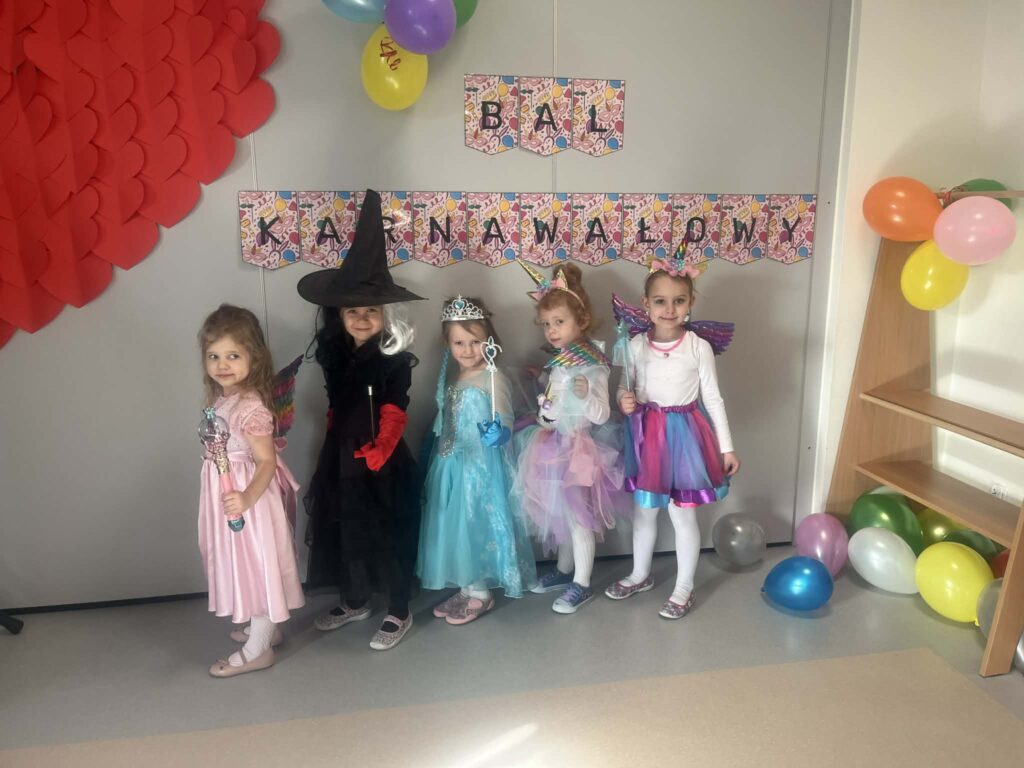 dziewczynki stoją przy napisie bal karnawałowy, dziewczynki są ubrane w stroje karnawałowe: czarownica i księżniczki