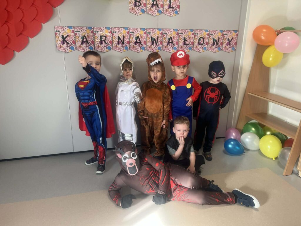 chłopcy stoją przy napisie bal karnawałowy, są przebrani w stroje karnawałowe: superman, kosmonauta, dinozaur, mario, spiderman, policjant i jeleń