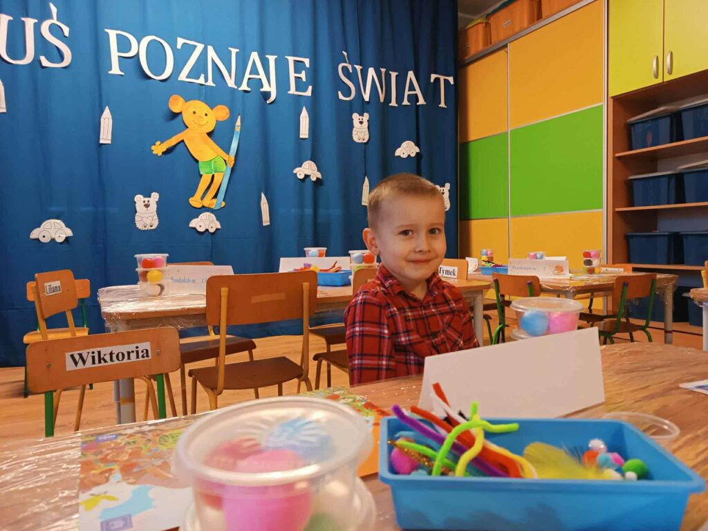 chłopiec w czerwonej koszuli, z tyłu plastuś z kredką, przed chłopcem nateriały plastyczne