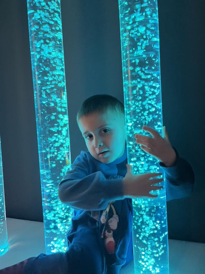 Chłopiec przytula się do sensorycznej tuby bąbelkowej. Obserwuje sekwencje kolorów, dotyka powierzchni kolumny wyczuwając wibracje.