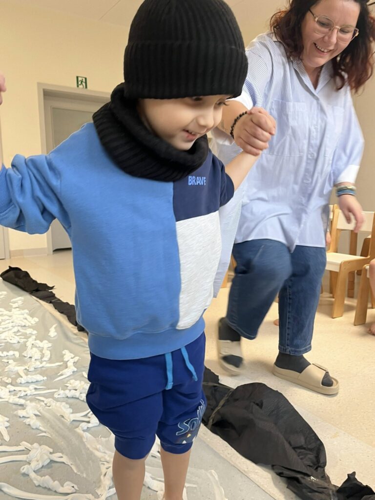 Chłopiec trzymany za ręce w celu asekuracji przez nauczycielki, ślizga się po stworzonym przez panie sztucznym lodowisku.