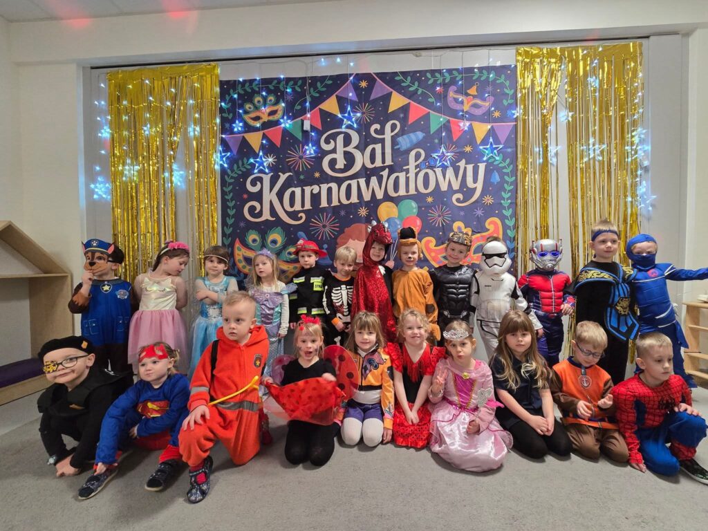 Duża grupa dzieci w kolorowych strojach karnawałowych pozuje do wspólnego zdjęcia na tle granatowej ściany ozdobionej serpentynami, maskami.