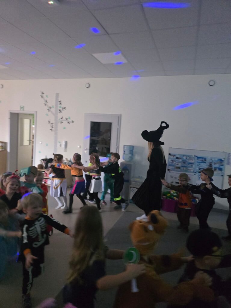 Duża grupa dzieci w kolorowych strojach karnawałowych bawią się na szarej sali i szarym dywanie. Tańczą do muzyki.