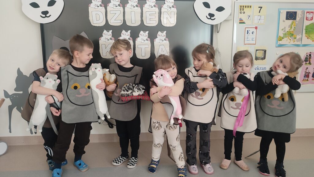 Czworo dzieci przebranych w stroje kotów pozuje do zdjęcia na tle napisu dzień Kota. Dzieci w rękach trzymają maskotkę kota, do której się przytulają.