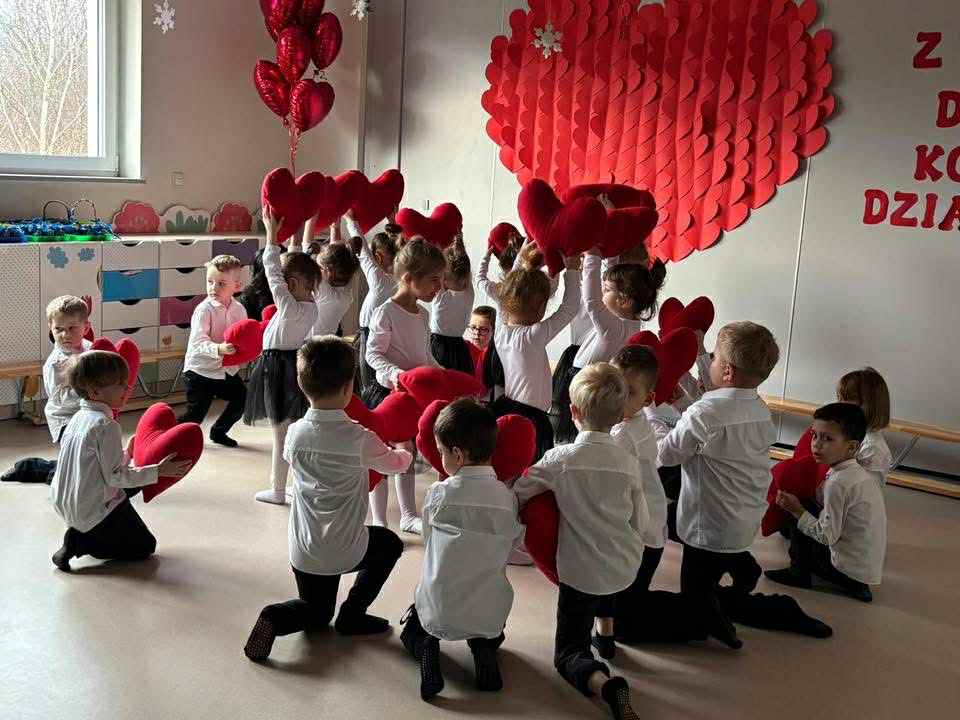 dziewczynki stoją po środku i trzymają serca w górze, chłopcy klęczą na zewnątrz i trzymają serca na dole