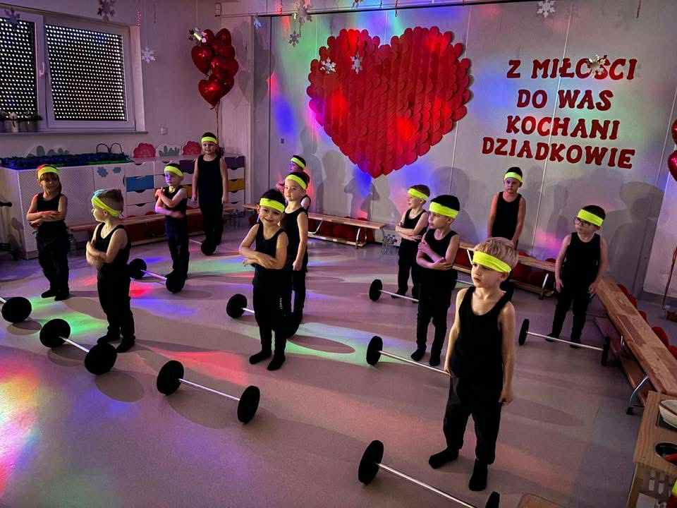 Chłopcy ubrani na czarno z jaskrawymi opaskami, stoją przed sztangami i przyjmują pozycje do tańca, dekoracja na ścianie duże serce i napis "z miłości do was kochani dziadkowie"