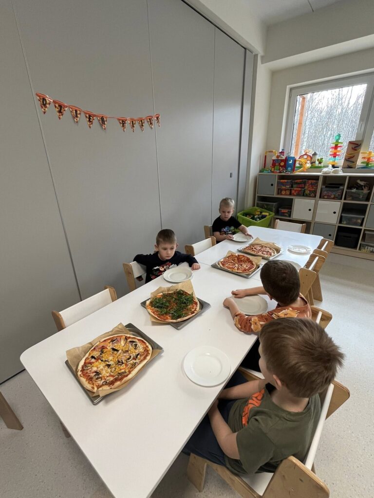 Dzieci siedzą przy stoliku. Jedzą pizzę zrobioną przez siebie.