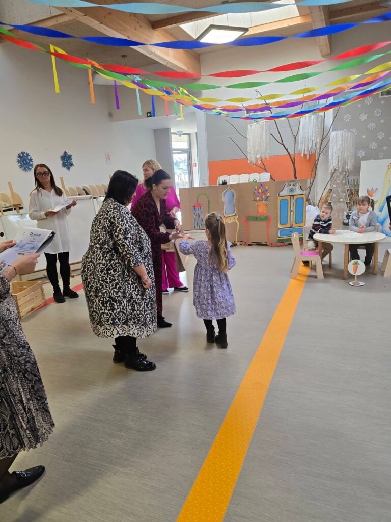 Dziewczynka w niebieskiej sukience odbiera nagrodę z rąk kobiety ubranej na czarno.