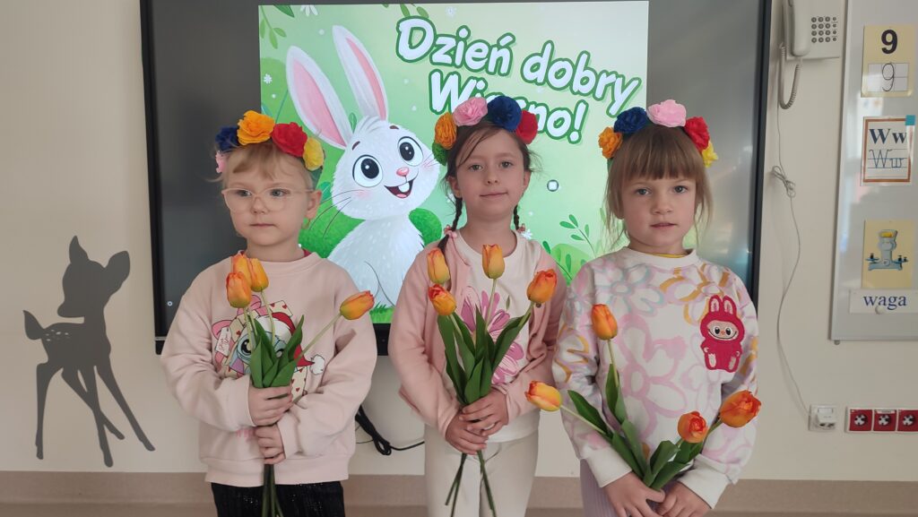 Trzy dziewczynki ubrane w kolory wiosny pozują do wspólnego zdjęcia. Za plecami obrazek wiosenny. Dzieci w rękach trzymają tulipany, na głowie mają wianki.