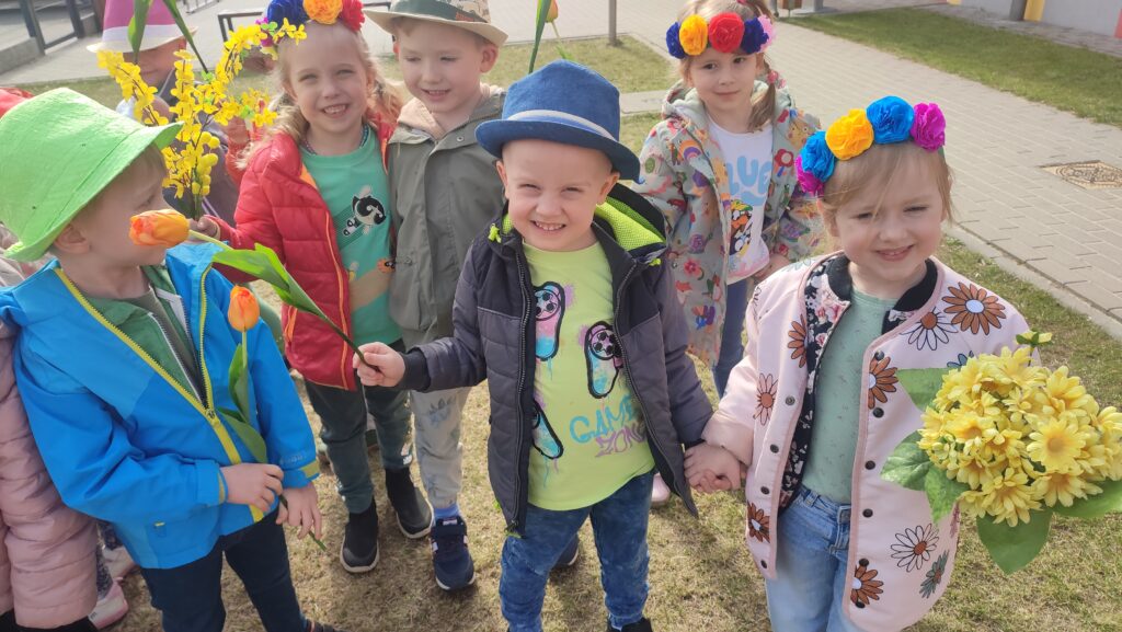 Duża grupa dzieci stoi na trawie w rękach trzymają atrybuty wiosny. Na głowach dzieci kolorowe kapelusze i kolorowe wianki.