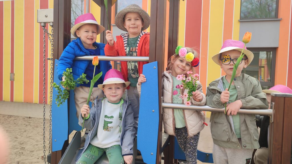Dziewczynki i chłopcy trzymają w ręce tulipany, forsycje, kwiatki. Pozują do zdjęcia na zjeżdżalni na placu zabaw.