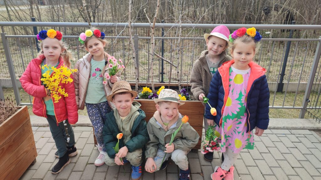 Chłopcy i dziewczynki ubrany w kolory wiosny, na głowach kapelusze i wianki, w rękach trzymają tulipany, forsycje, kwiatki. Pozują na tle drzew wiosennych.