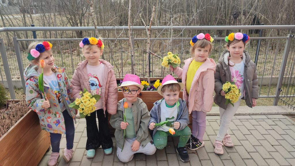 Chłopcy i dziewczynki ubrany w kolory wiosny, na głowach kapelusze i wianki, w rękach trzymają tulipany, forsycje, kwiatki. Pozują na tle drzew wiosennych.