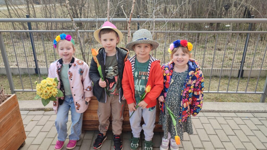 Chłopcy i dziewczynki ubrany w kolory wiosny, na głowach kapelusze i wianki, w rękach trzymają tulipany, forsycje, kwiatki. Pozują na tle drzew wiosennych.