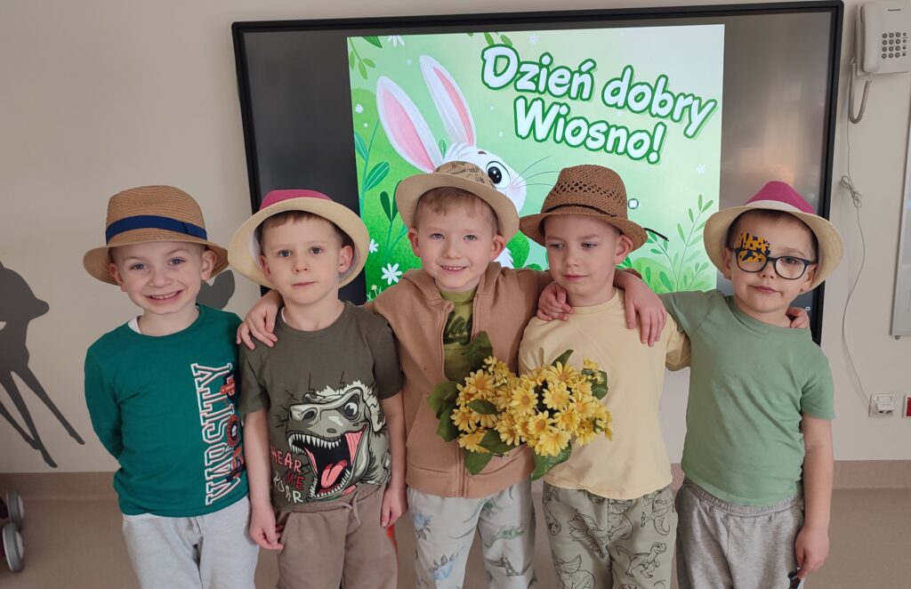 Pięć chłopców ubranych w kolory wiosny pozują do wspólnego zdjęcia. Za plecami obrazek wiosenny. Dzieci w rękach trzymają kwiatki, na głowie mają kapelusze.