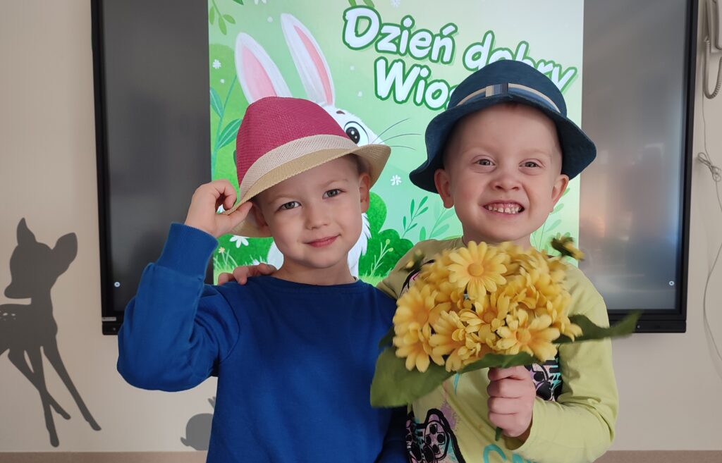 Dwoje chłopców ubranych w kolory wiosny pozują do wspólnego zdjęcia. Za plecami obrazek wiosenny. Dzieci w rękach trzymają kwiatki, na głowie mają kapelusze.