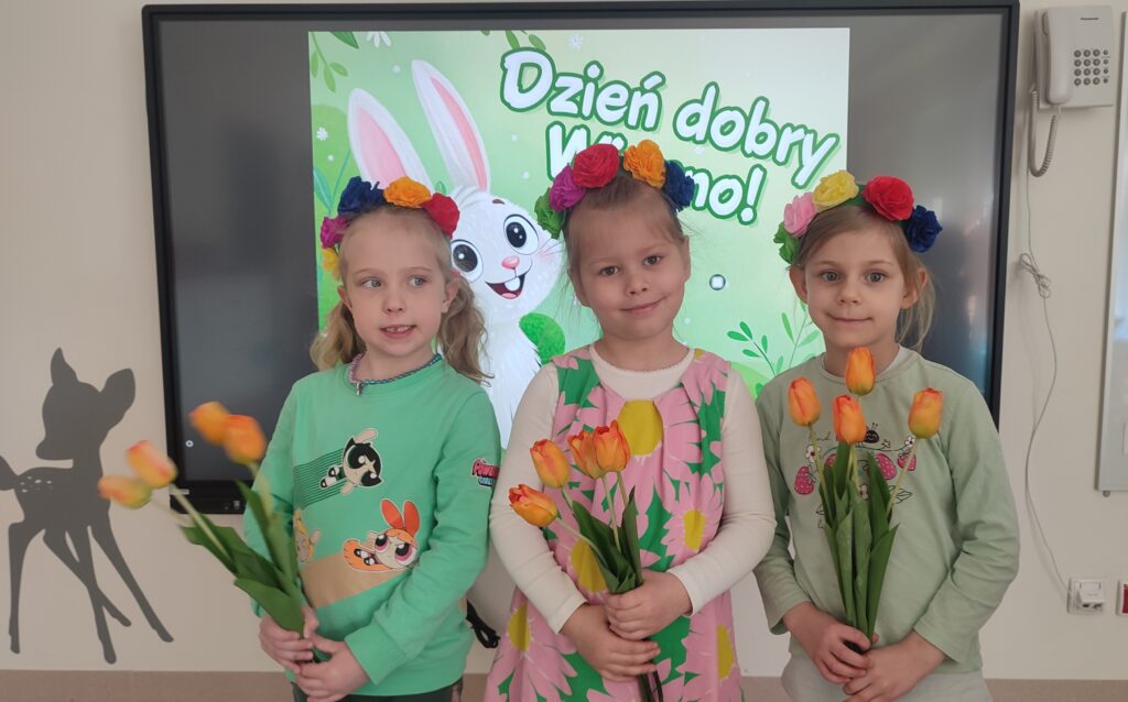 Trzy dziewczynki ubrane w kolory wiosny pozują do wspólnego zdjęcia. Za plecami obrazek wiosenny. Dzieci w rękach trzymają tulipany, na głowie mają wianki.