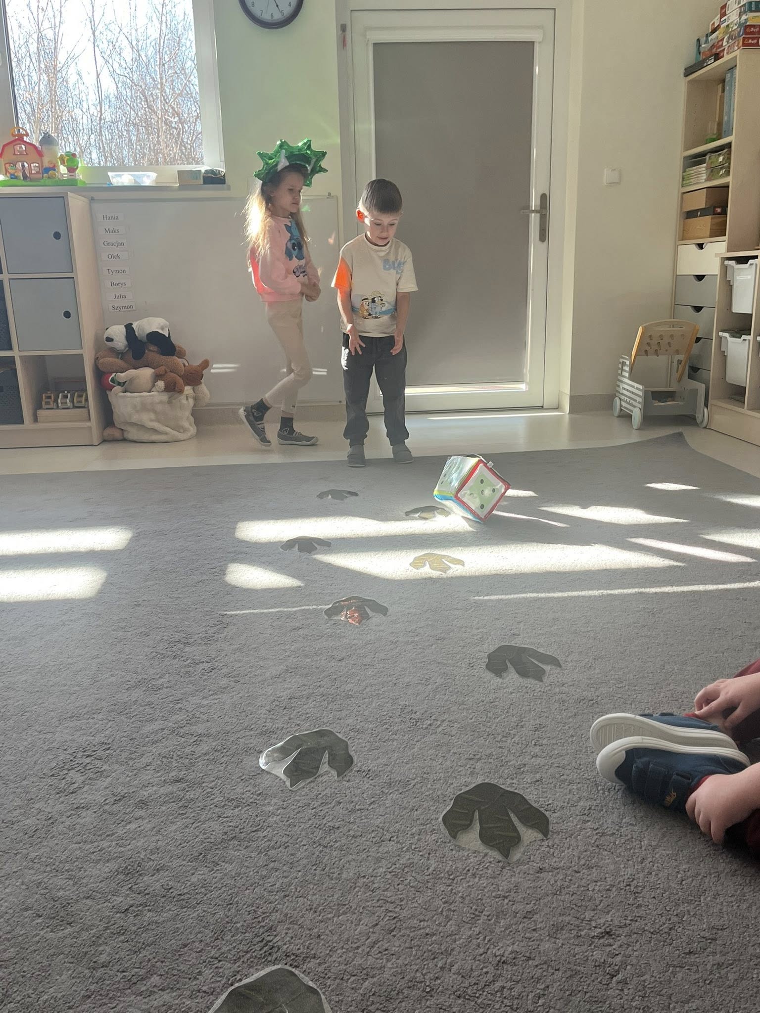 Chłopiec z dziewczynką skaczą po śladach dinozaura zgodnie z wyrzuconą na kostce liczbą oczek.