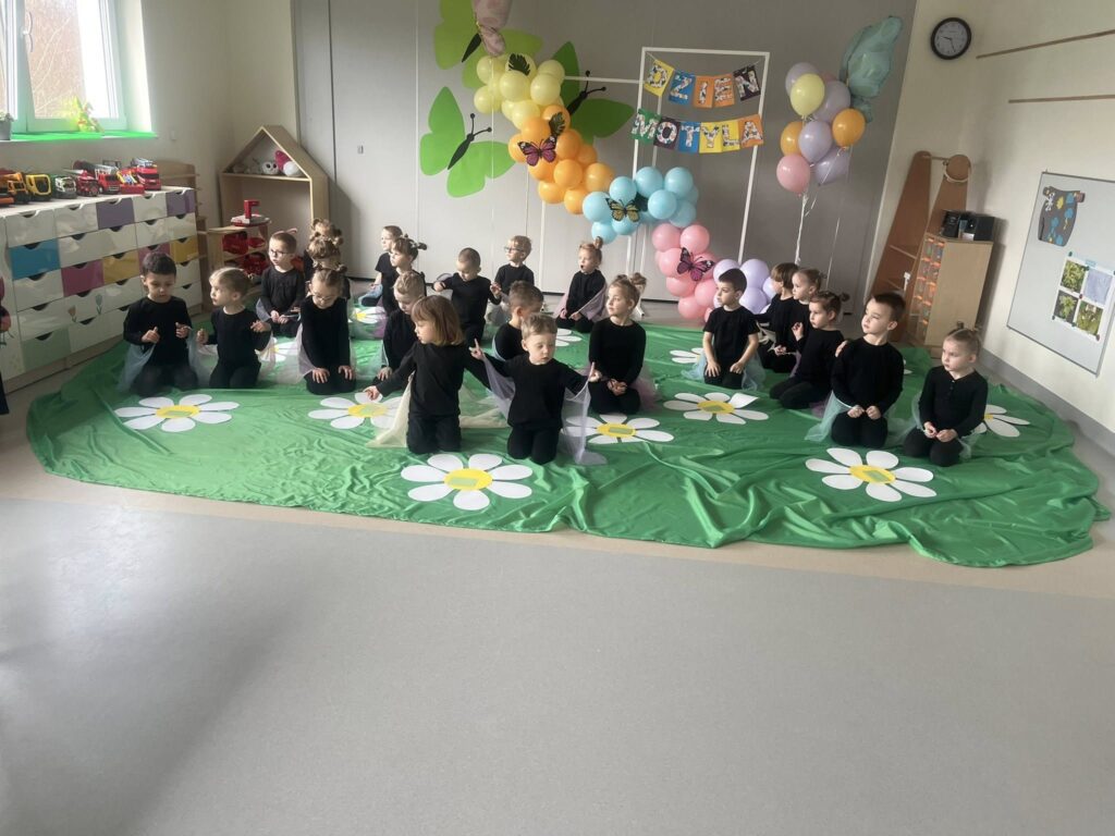Dzieci siedzą w strojach motyli, na zielonym materiale przy białych kwiatkach