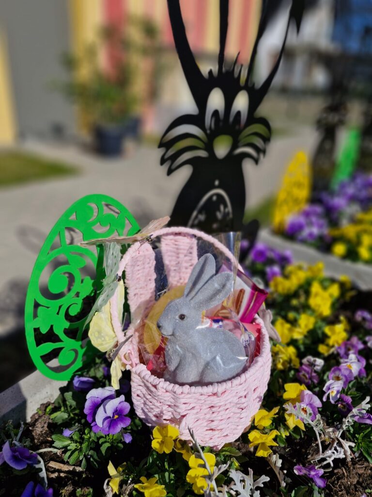 Biały koszyczek wielkanocny wypełniony prezentami stoi wśród kolorowych kwiatków. Obok duże pisanki i zajączek.
