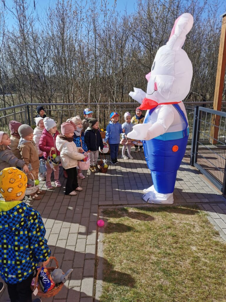 Dzieci obok budynku przedszkola witają wielkanocnego Zajączka