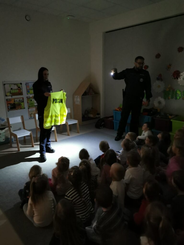 Policjant świeci latarką na kamizelkę odblaskową., dzieci obserwują.