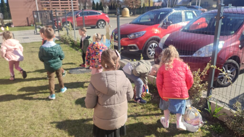 Grupka dzieci na trawie szuka ukrytych koszyczków wielkanocnych.