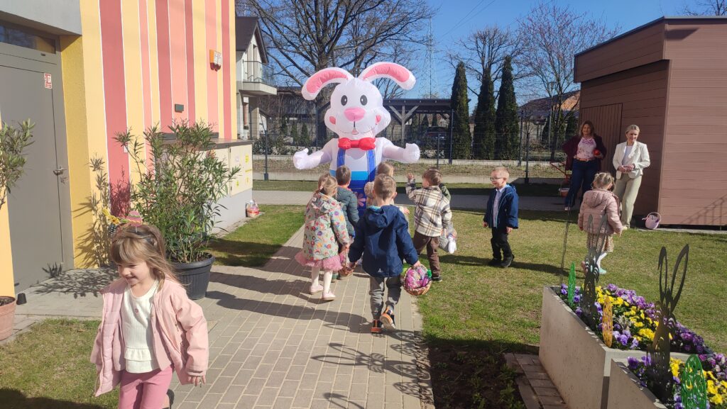 Grupka dzieci z koszyczkami wielkanocnymi idzie po chodniku do dużego zajączka wielkanocnego, który jest biały.
