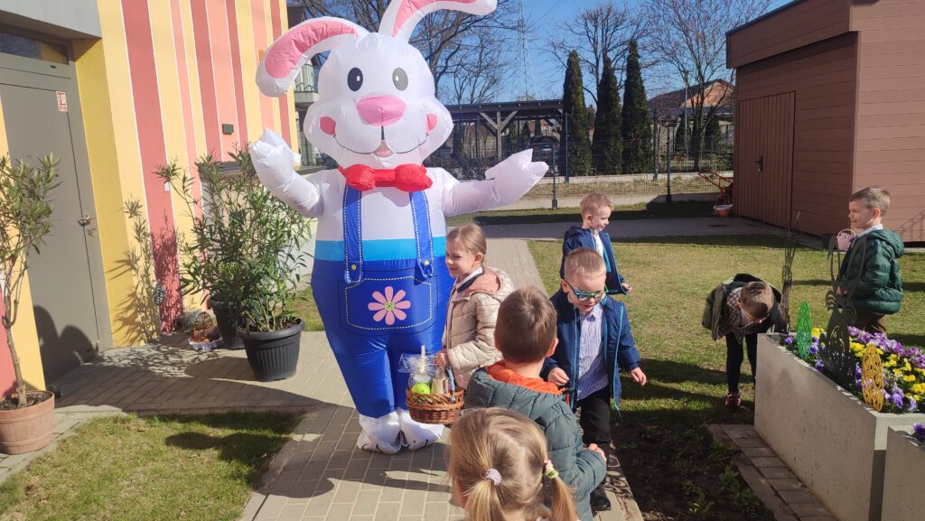 Grupka dzieci z koszyczkami wielkanocnymi idzie po chodniku do dużego zajączka wielkanocnego, który jest biały.