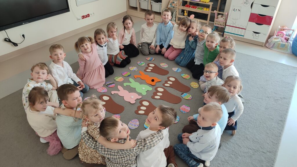 Duża grupa dzieci siedzi na szarym dywanie w dużym kole. W środku leżą sylwety kolorowych zajączków, jego duże łapy i kolorowe pisanki.