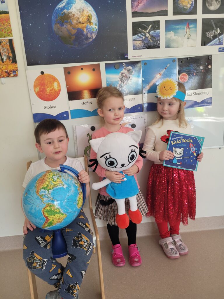 Trójka dzieci stoi pod tablicą trzymając globus, Kicię Kocię i książkę
