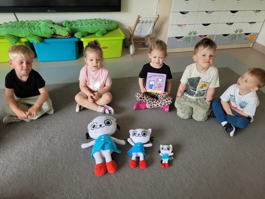 Trójka dzieci siedzi na dywanie. przed nimi leżą trzy maskotki Kici Koci