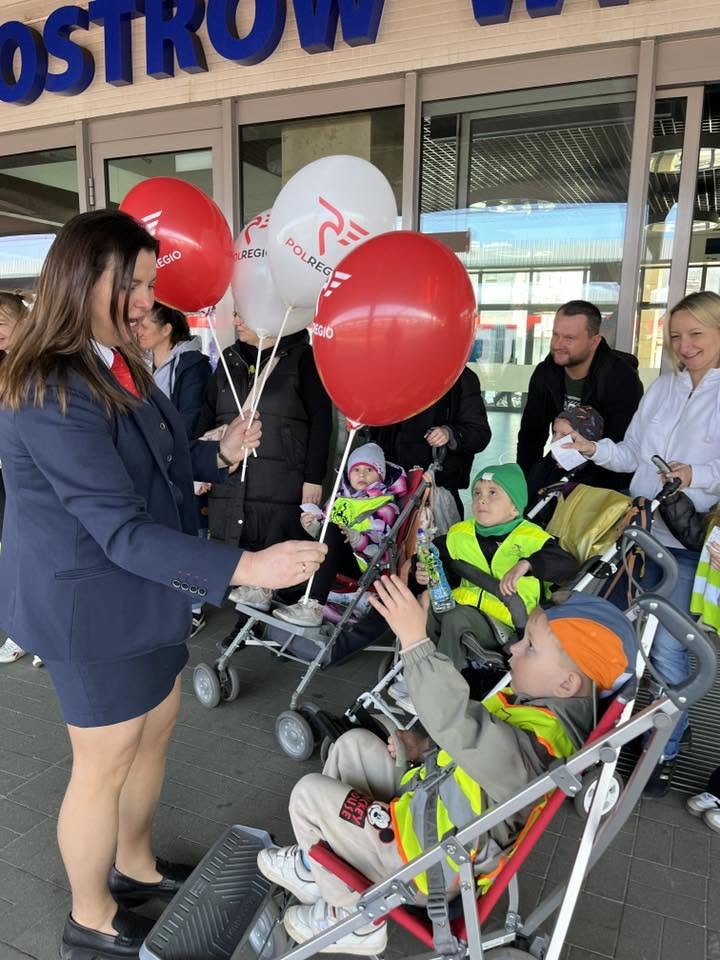 Dzieci stoją przed dworcem kolejowym i otrzymują od Pani Konduktor czerwone i białe balony.