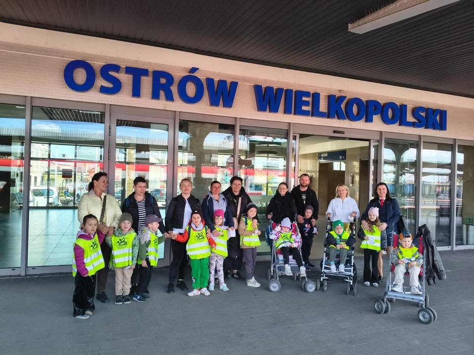 Dzieci wraz z opiekunami stoją przed dworcem kolejowym i pozują do zdjęcia.