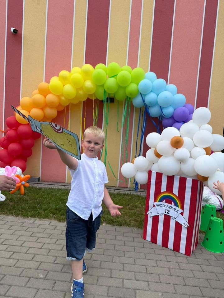 Chłopiec ubrany w białą koszukę i krótkie spodenki pozuje do zdjęcia na tle kolorowej balonowej tęczy.