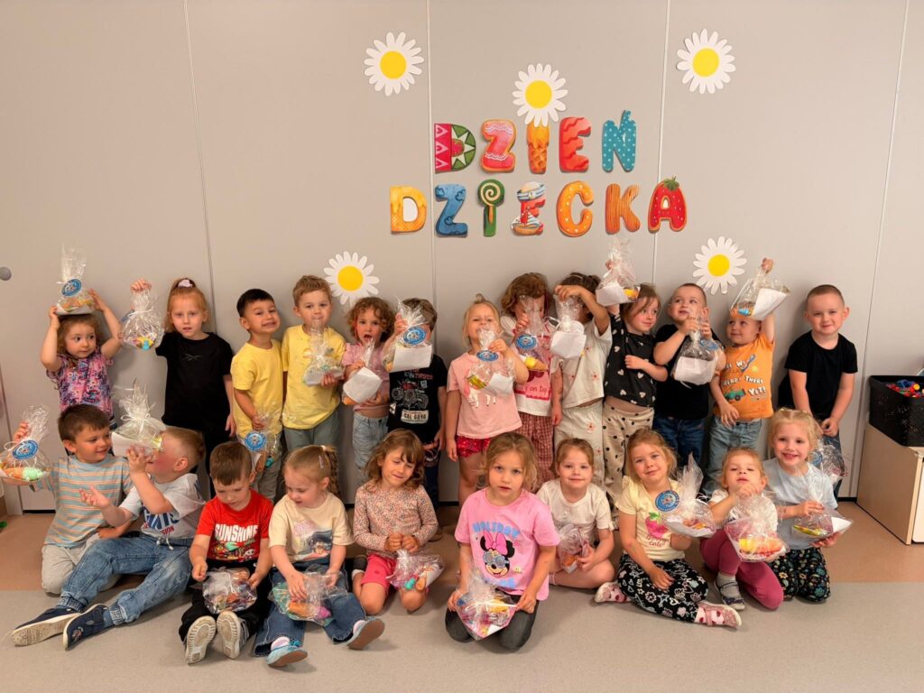 Na zdjęciu widoczna jest grupa dzieci którzy stoją na tle ściany z napisem "Dzień Dziecka" oraz widocznymi kwiatuszkami. Dzieci w rączkach trzymają prezenciki.