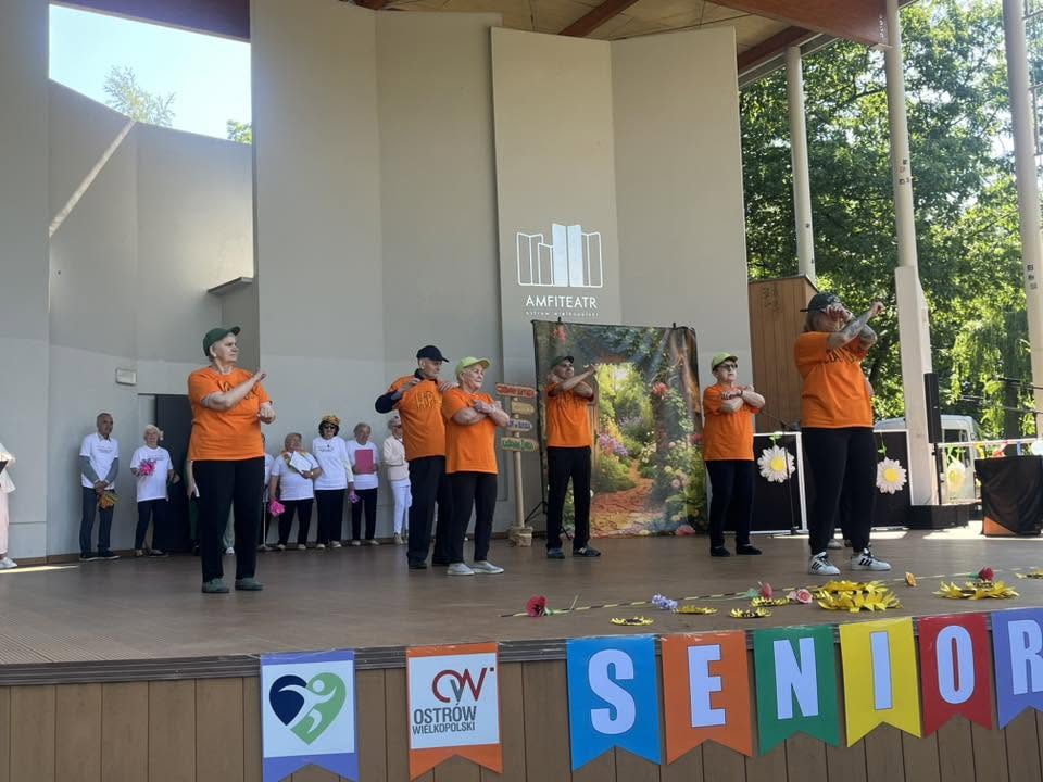 seniorzy w pomarańczowych koszulkach stoją na scenie z założonymi rękami 
