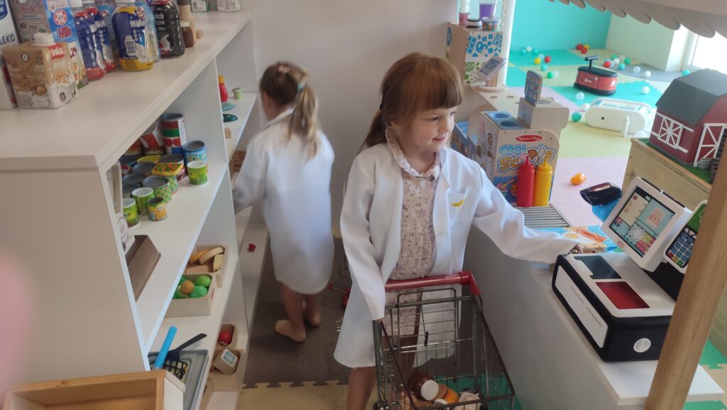 Dwie dziewczynki ubrane w białe fartuchy kupują produkty spożywcze w dużym drewnianym sklepie zabawkowym.